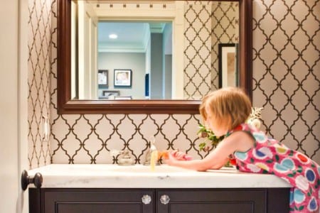Little girl using the sink in in bathroom. Bathroom wallpaper is a black and white pattern.