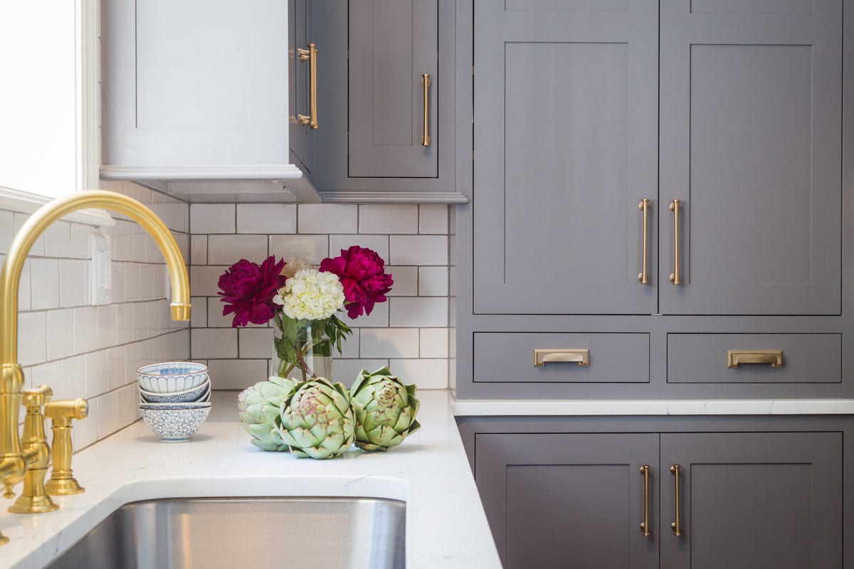 close up of a kitchen with white backsplash and grey cabinetry with gold fixtures