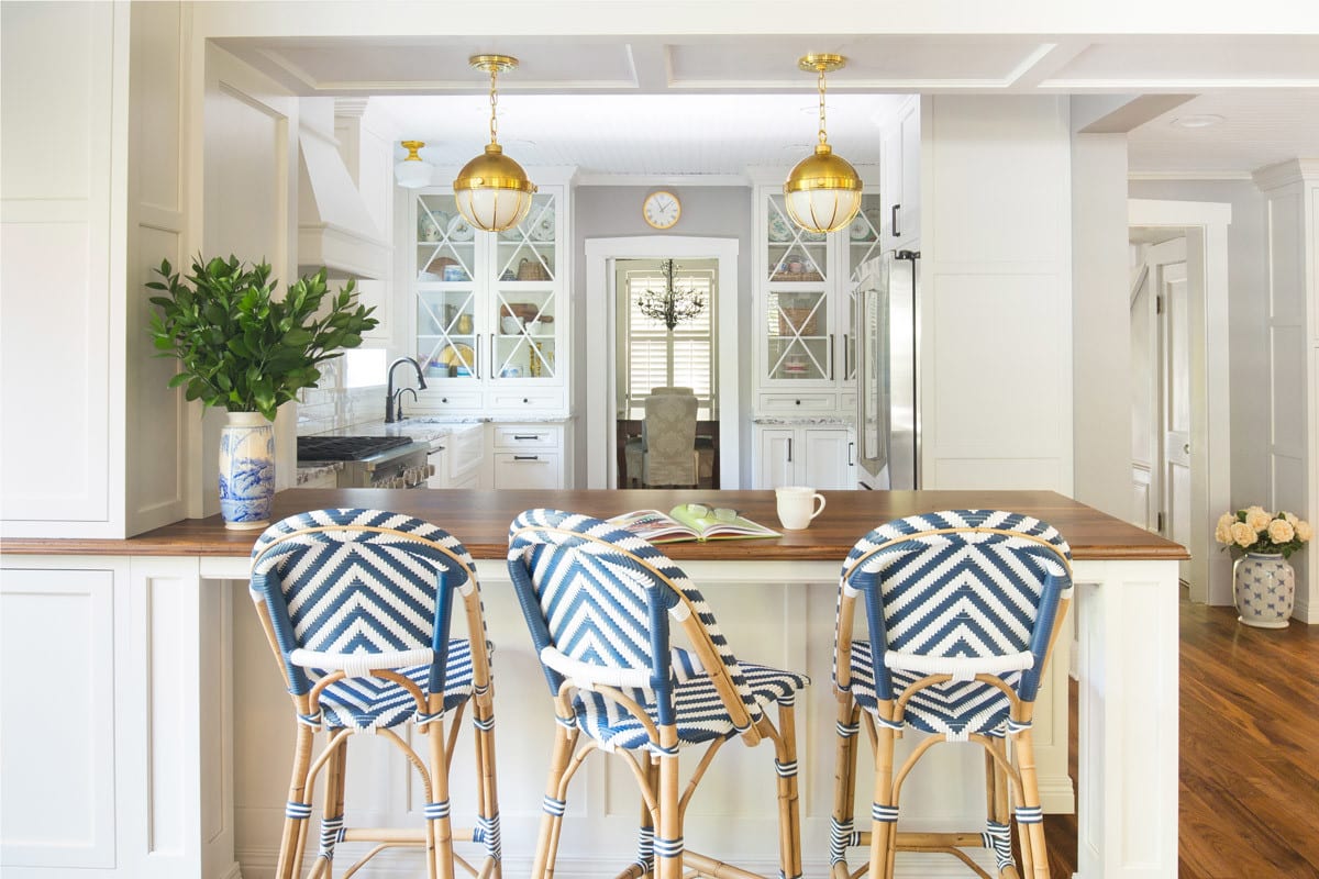 Two symmetrical cabinets that sit on the counters with glass X mullions were designed to showcase the homeowner’s beautiful dishes and offer an added layer of charm for the kitchen. A peninsula was added to the kitchen with unique brass pendants that hang from the soffit and hang in perfect symmetry for line of sight into the kitchen. The cabinets are a soft white and the island has a distressed walnut thick top to accent the white farmhouse shaker sink. And who can forget the pop of color navy counter stools which are fun and youthful.