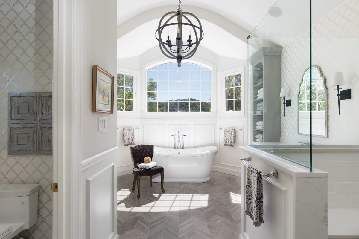 large white bathroom remodel with tub, shower, toilet, brown chair, tall grey cabinet, large white window for natural light. gold mirror on white grid wall with large grey sphere chandelier.
