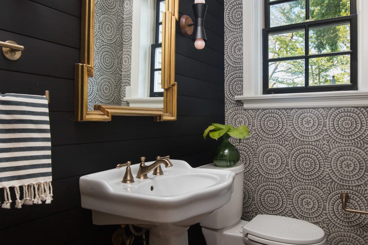 black wall with golden mirror and bathroom white sink and toilet. green plant by window for natural light on swirly grey and white wall bathroom remodel design