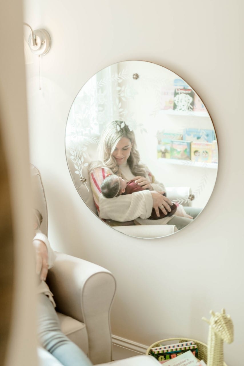 A mirror reflects an image of a woman sitting holding a child in her lap in a chair in a corner of a room, bathed in natural light from the windows surrounding them.