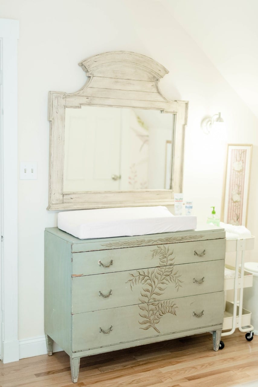 A rustic pastel-colored changing table with a mirror on top of a set of drawers next to a rolling cart with supplies.