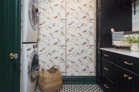 A dark green, black, and white laundry room with a washer and dryer by bird-patterned wallpaper. The black shelves and cabinets have golden handles and house necessary laundry products.