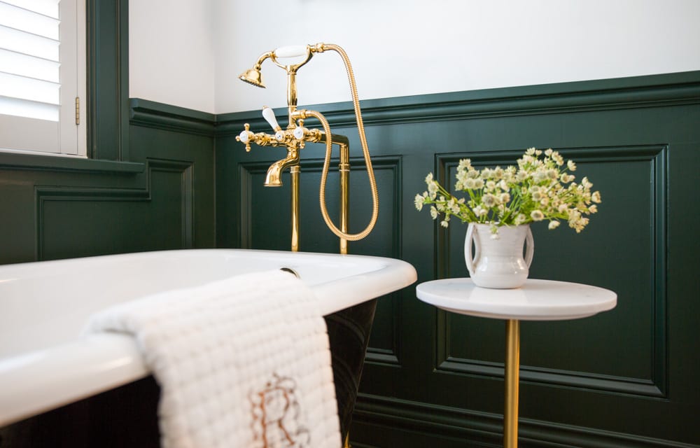 A small plant sits atop a nightstand next to a bathtub in a primarily dark green-and-white bathroom with golden accent pieces.