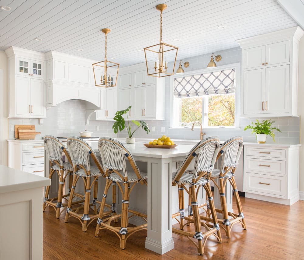 A white kitchen with wooden floors and chairs. A window lets in natural light that reflects off of golden light fixtures.