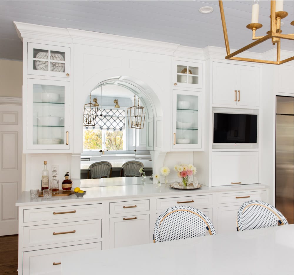 White cabinets with a mirror in the center behind a kitchen island with blue white and brown chairs