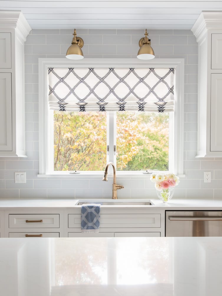 White kitchen with hints of navy blue present in the curtains and dishtowel and natural light entering from a window above the sink.
