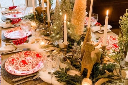 A large winter holiday table decoration spread with red decorative plates, tall wine glasses, and foliage and candles on the table runner.