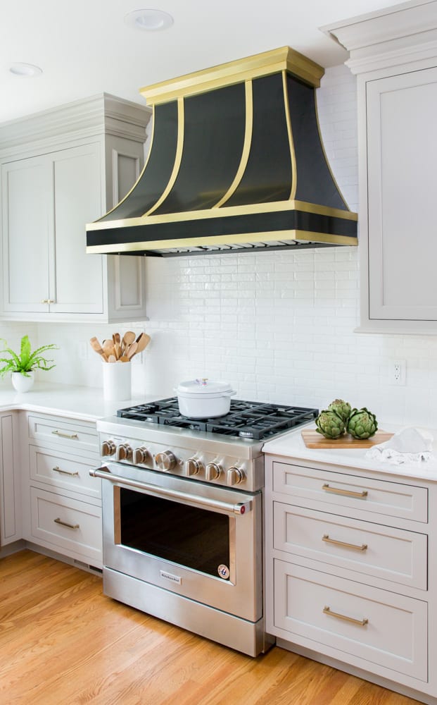 A kitchen with a white brick backsplash and light grey cabinets along with a large black-and-gold vent fan above a silver stove.