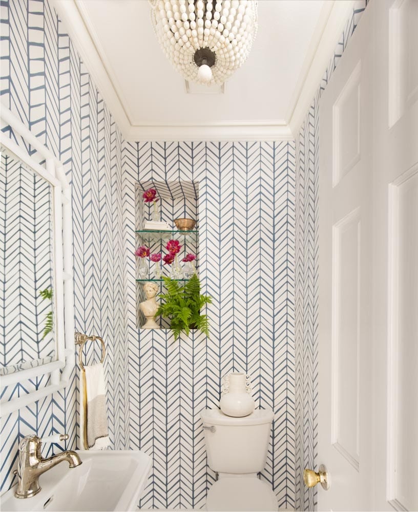 A great gatsby-styled bathroom with herringbone-patterned wallpaper, white sink, toilet, and door, and golden accents