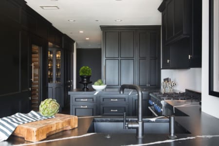 Black kitchen with white ceilings and walls with silver handles and accents and a wooden cutting board next to the sink.