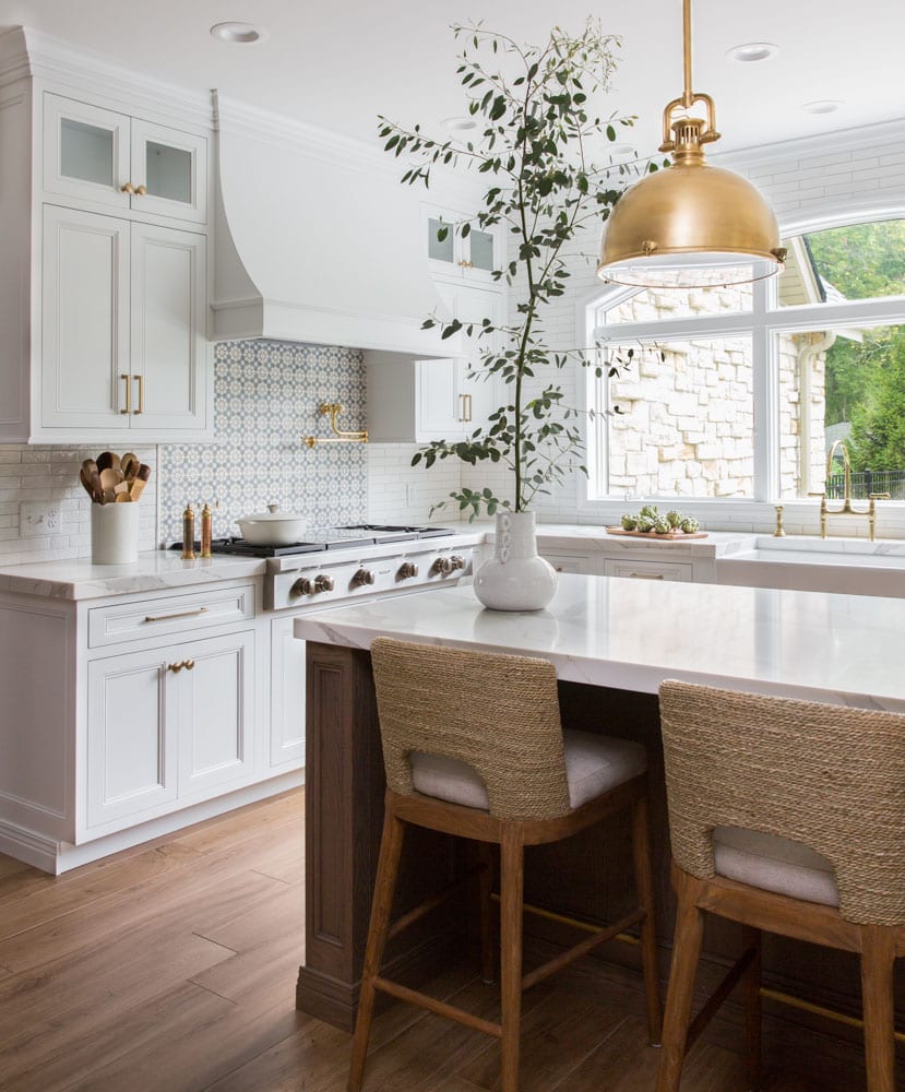 A white kitchen with a large window letting in natural light and an island table in the center surrounded by chairs.
