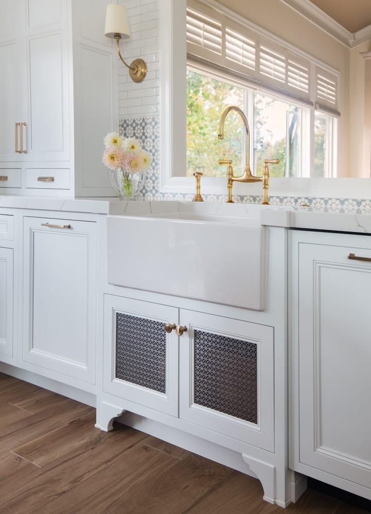 A white kitchen sink sits atop white cabinets with a large mirror behind it, which reflects a large white window letting in natural light.