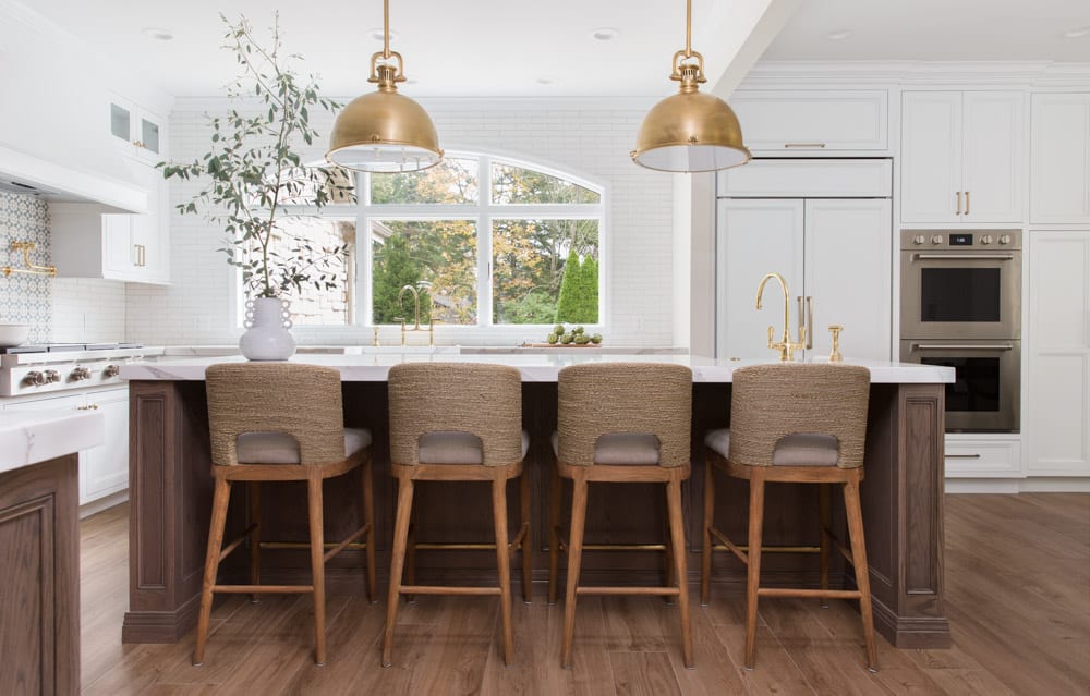 A large kitchen with white walls, cabinets, and countertops, along with wooden floors and chairs. Accents of gold are present in faucets, handles, and hanging light fixtures.