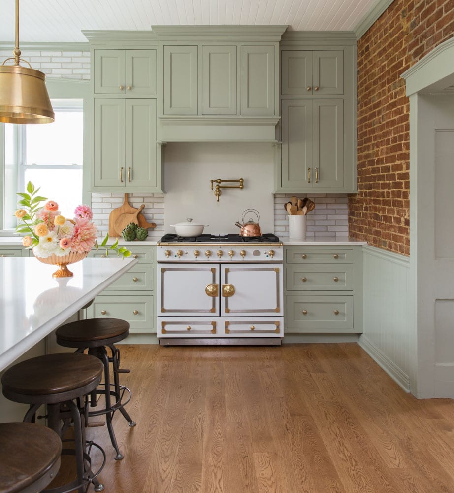 Pastel green cabinets surround a white and gold oven and stove next to a kitchen island countertop with brown stools beneath it.