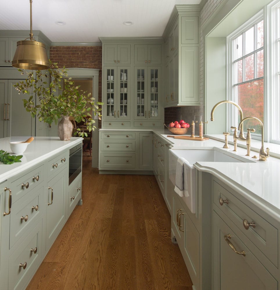 Pastel green cabinets surround a small, but long, kitchen with white countertops and golden handles, faucets, and light fixtures.
