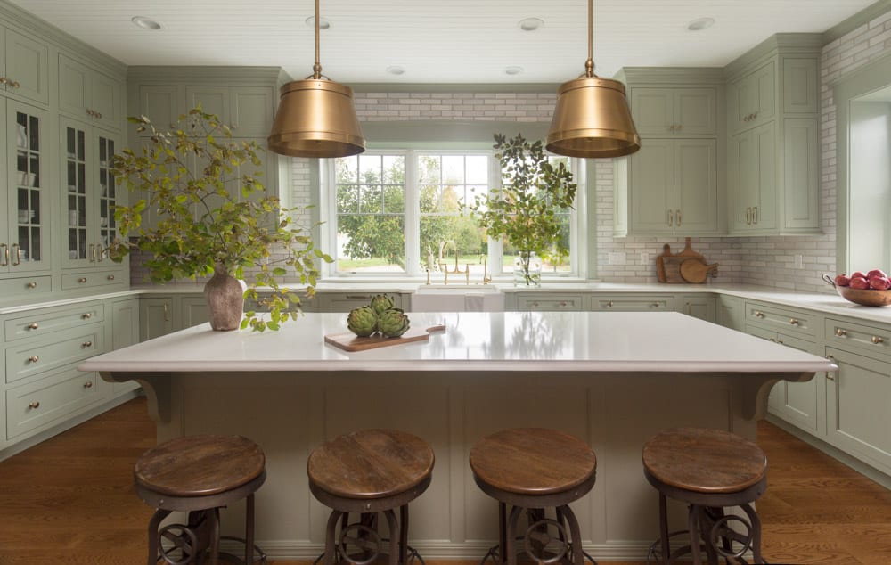 A pastel green kitchen with white brick backsplash and white countertops. There are brown stools at the island table in the center and the only light is natural light from a large window above a golden sink.