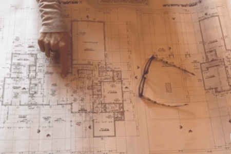 Hand pointing at a floor plan of a house with glasses sitting on top of the paper.