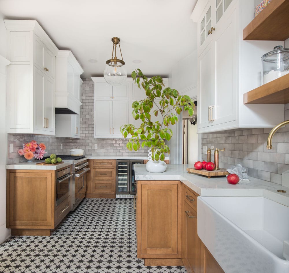 Long kitchen with white and wooden cabinets and a black and white-tiled flooring. A plant and some fruits add accents of green and red.