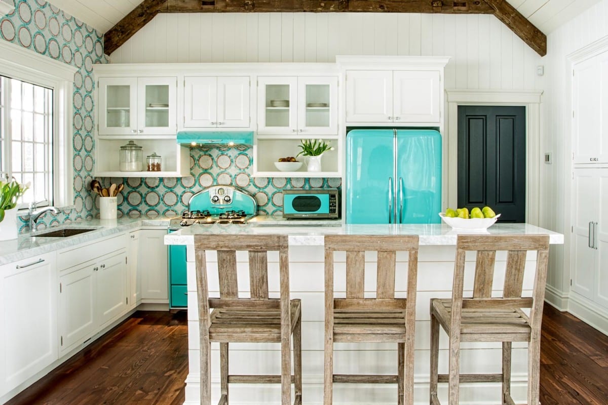 White kitchen interior design remodel with brown wooden floor, trim, and chairs, with bright teal accents. Teal retro fridge, oven, microwave, and backsplash. White floating cabinets and island bar with green plants and wooden kitchen utensils.