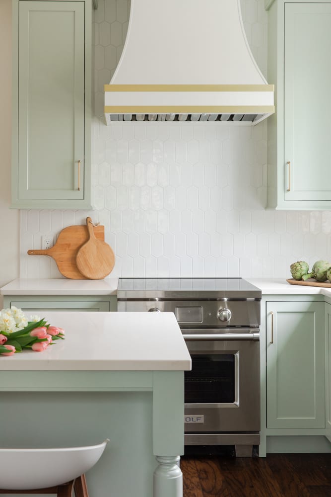 The cabinets are sage green with gold hardware and the countertops are white. The stove hood is white and gold