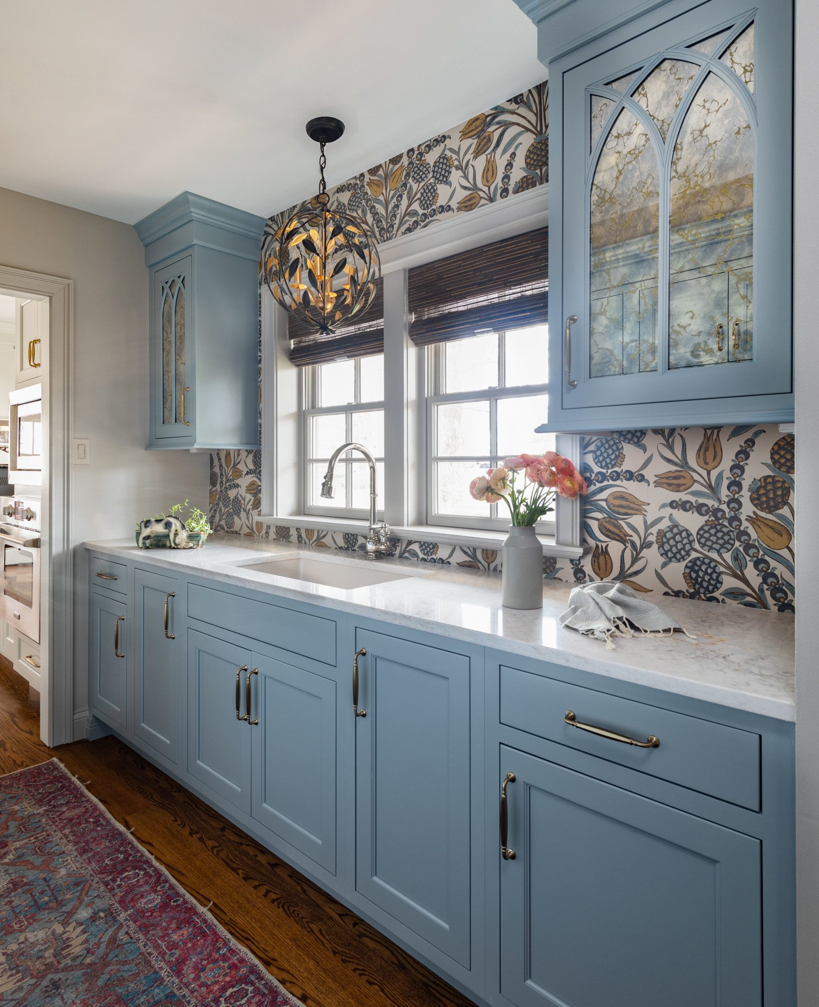 Kitchen cabinets are blue and the walls are patterned with intricate wallpaper and a gorgeous light fixture above the sink.