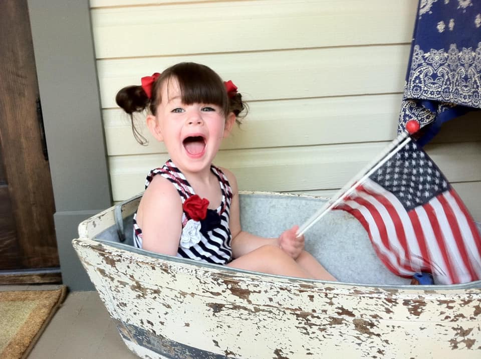 A cute baby with pigtails laughing inside of a vintage washbasin, holding flags