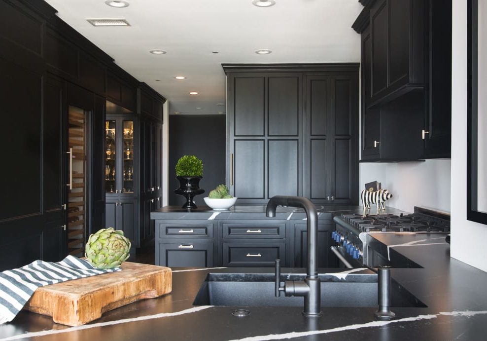 Black kitchen with white ceilings and walls with silver handles and accents and a wooden cutting board next to the sink.