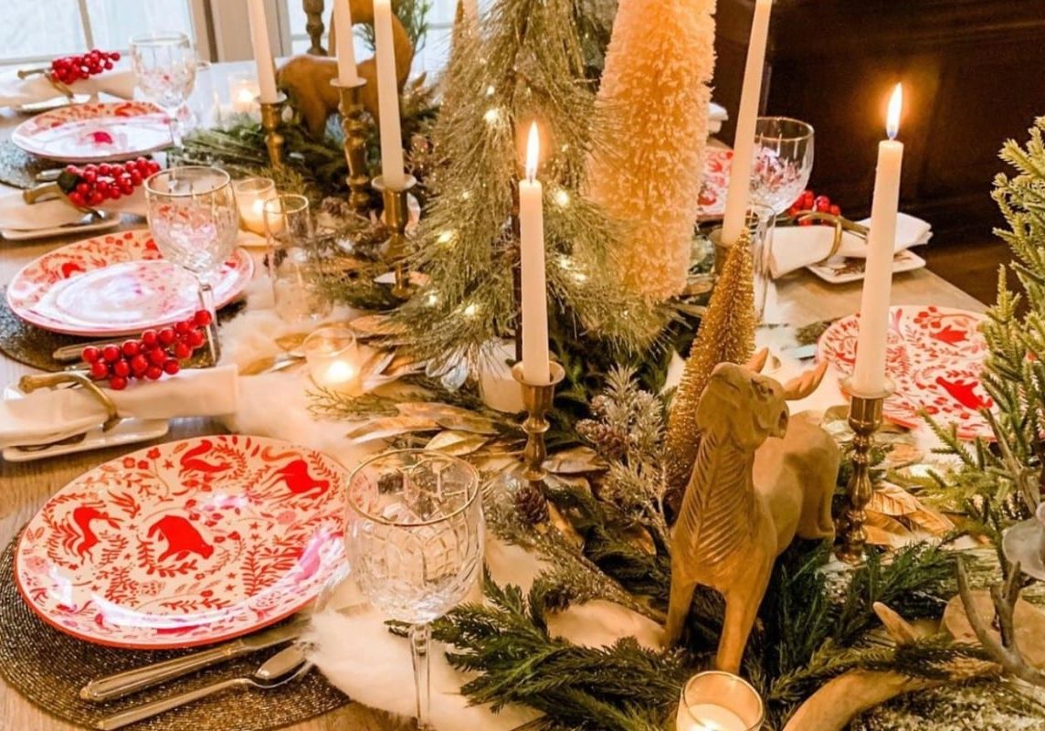 A large winter holiday table decoration spread with red decorative plates, tall wine glasses, and foliage and candles on the table runner.