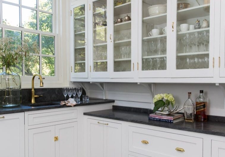 Lindell-282200930 A kitchen sink sits under a window next to large glass cabinets filled with glasses and dishes for serving food and drinks.