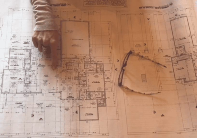 Screen Shot 2021-01-15 at 2.56.50 PM Hand pointing at a floor plan of a house with glasses sitting on top of the paper.
