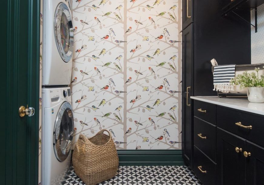 A dark green, black, and white laundry room with a washer and dryer by bird-patterned wallpaper. The black shelves and cabinets have golden handles and house necessary laundry products.