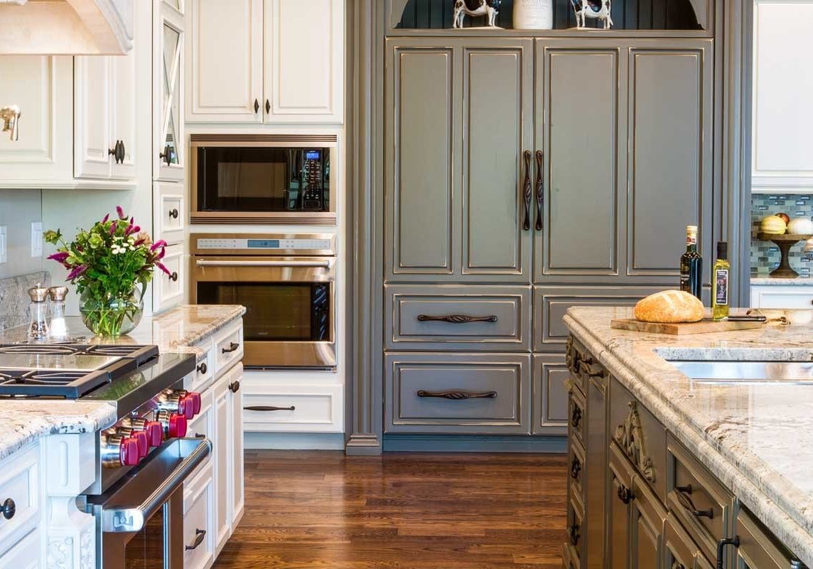Kitchen cabinets are white and the island cabinets are a weathered grey. The fridge is hidden behind large grey cabinets that match the island.