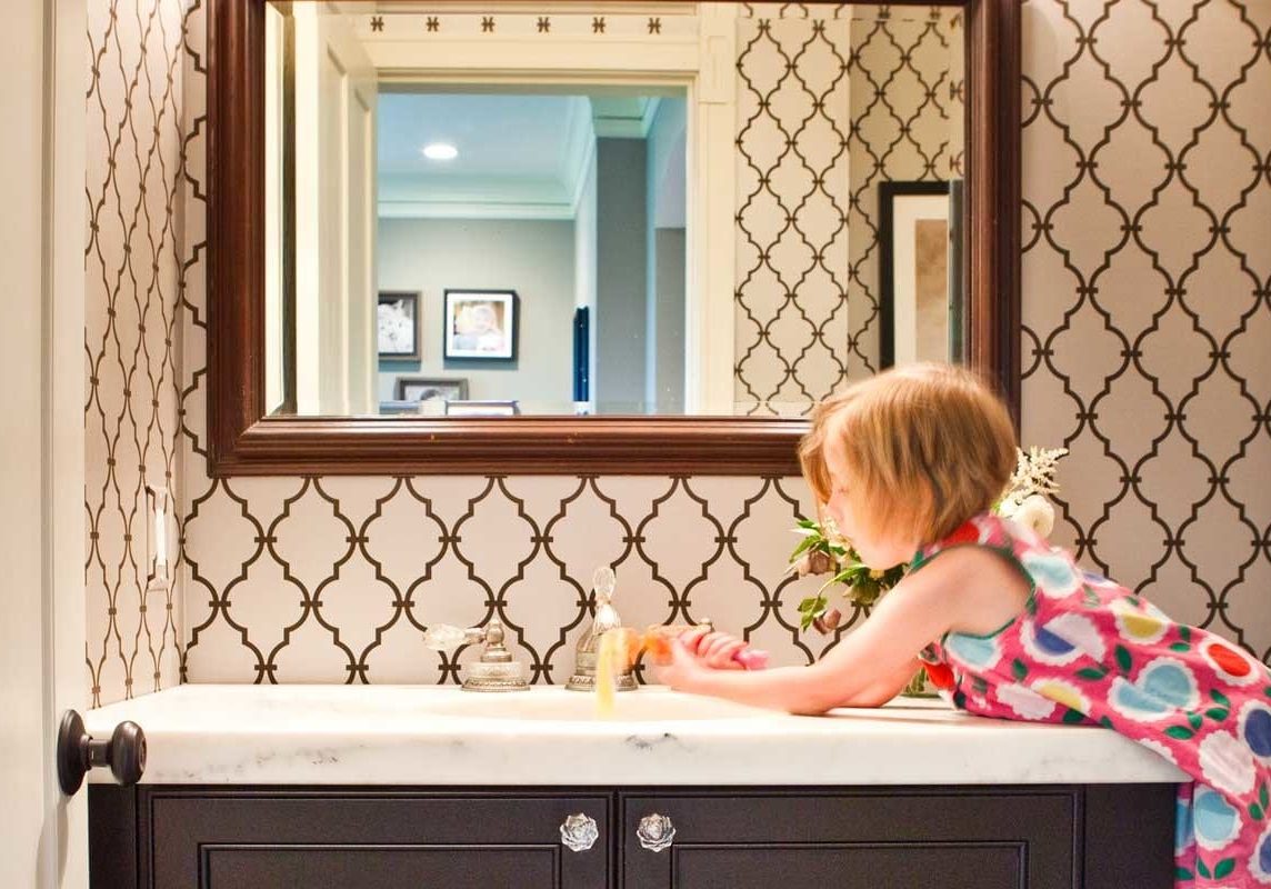 Little girl using the sink in in bathroom. Bathroom wallpaper is a black and white pattern.